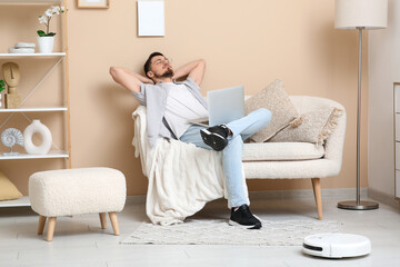 Modern robot vacuum cleaner near sofa with resting young man at home