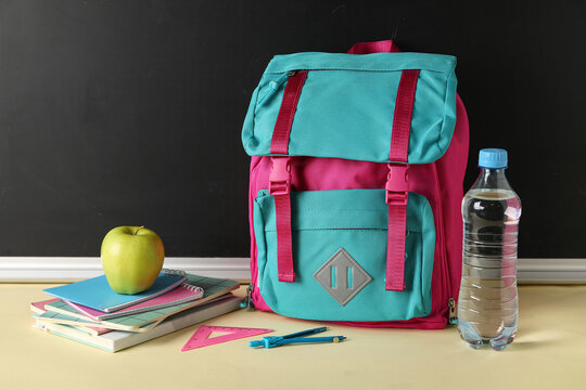 School backpack, stationery, bottle of water and apple on table against chalkboard