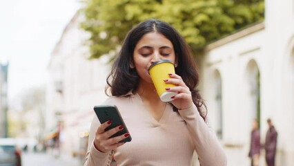 Indian woman using smartphone typing text answering messages chatting online looking mobile screen social media app outdoors. Girl enjoying morning coffee drink on urban city street. Town lifestyles