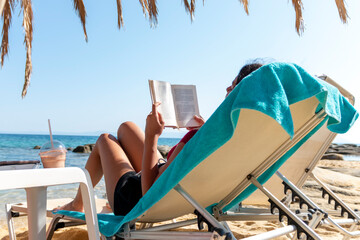 A young woman with long hair is sitting on a beach lounge. She is deeply engrossed in a captivating book, completely absorbed in the story as she enjoys the peaceful ambiance of the beach.