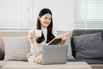 A woman is sitting on a couch with a laptop and a cup of coffee. She is reading a book while sipping her coffee. Concept of relaxation and leisure, as the woman enjoys her time reading