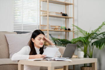 A woman is sitting on a couch with a laptop and a stack of papers in front of her. She is looking at the papers and she is in a state of distress