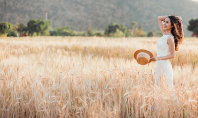 A woman stands in a field of tall golden wheat, holding a straw hat. The scene is serene and...