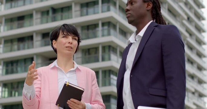Woman real estate agent talking to male client against backdrop of modern buildings. Concept of property consultation, urban real estate, and professional communication.