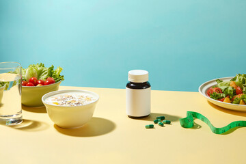 Losing weight theme photography with fruit yogurt, lettuce, cherry tomatoes, water, vegetable salad and a drug bottle without label next to some green pills flat lay on table. Front view, copy space