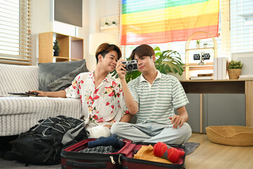 Smiling homosexual couple sitting on floor in living room and packing clothes into travel bag