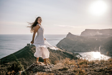 A woman stands on a hill overlooking the ocean. She is wearing a white dress and has long hair. The...