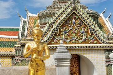 Gold chimera statue in front of a decorative side gate between the Temple of the Emerald Buddha (Wat Phra Kaew) and the Grand Palace in Phra Nakhon, Bangkok, Thailand on a fine day with blue skies