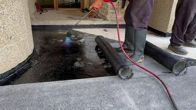 Construction worker heating with a blow torch an bituminous material and rolled across a concrete surface