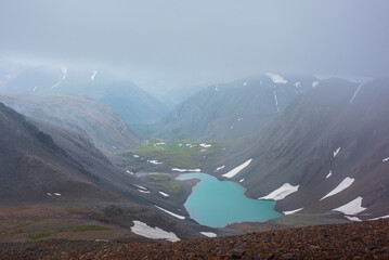 Most beautiful turquoise alpine lake in stony rocky green valley against mountain range silhouette in rainy low clouds in gray sky. Large snowy mountains in light transparent mist in rain bad weather. - Powered by Adobe