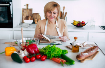 Close up portrait of beautiful smiling mature woman in apron is reading a book with recipes while cooking in the modern light kitchen with many vegetables on the table