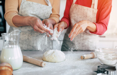 Close up image of hands of mother and daughter spreading wheat flour on the dough and preparing pastries in the kitchen at home