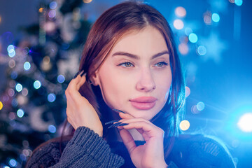 Close-up portrait of a young attractive woman dressed in a sweater posing against the background of a Christmas tree.