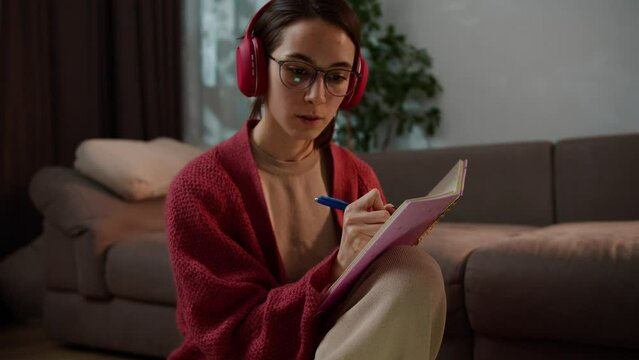 Close-up of a confident brunette girl with glasses in a red sweater and wireless headphones studying foreign languages online and taking notes in her notebook in a modern apartment