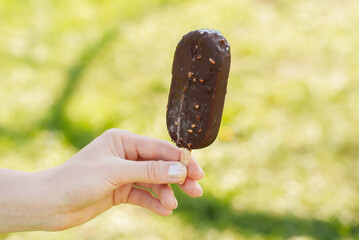 Ice cream on stick in chocolate glaze with pieces of nuts in female hands on natural background,...