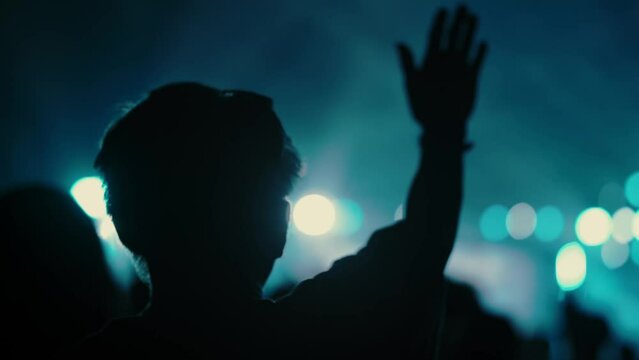 "In a slow-motion shot, a man raises his hands in reverence, praising and singing worship songs at a Christian festival. The scene is set with a large crowd, a stage illuminated with vibrant lights an