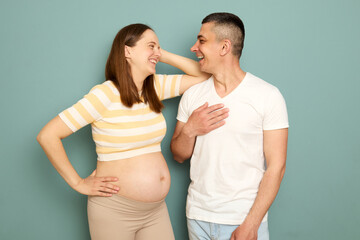 Satisfied happy pregnant couple looking at each other with love and smiles woman with bare belly leaning on her husband shoulder isolated over light green background