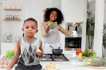 African boy child eat snack while blurred background of curly hair mother hold fresh vegetable...