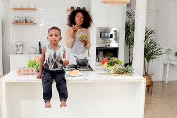 African boy child eat snack while blurred background of curly hair mother hold fresh vegetable salad bowl at kitchen, mom and son prepare meal together. Beautiful family cooking healthy food at home.