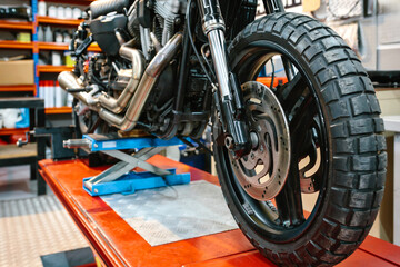 Close up of front wheel tyre and brake disk of custom motorbike on workshop. Front view of vintage scrambler motorcycle over platform ready to repair on cleaned mechanic garage. © David Pereiras
