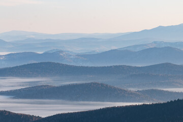 Aerial perspective in the mountains with peaks overgrown with forest and fog between them stretching into the distance to the horizon