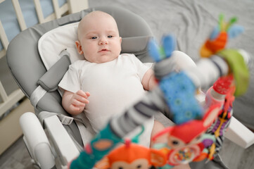 Serious newborn lies in cocoon chaise lounge, baby carefully examines multi-colored toys on...