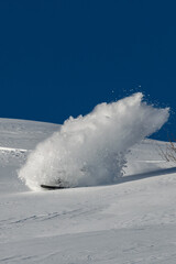 A freerider snowboarder lifts a huge cloud of snow while making a turn in deep snow against the backdrop of a blue sky and an untouched slope