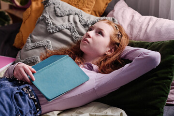 Teen girl holding book relaxing on bed in her bedroom thinking about plot of novel she reading,...