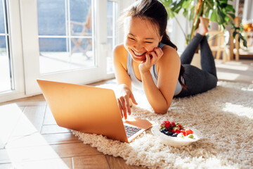 Young Asian woman lies on carpet in living room and uses laptop. Attractive female Korean is...