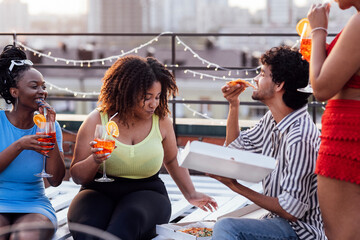 Four cheerful friends are eating pizza and enjoying delicious cocktails. Roof party. Dark-skinned...