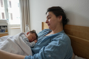 Obraz premium mother holds her newborn baby close to her chest in bed, exuding warmth and tenderness. The baby sleeps soundly, wrapped in a blanket, while the mother gazes lovingly, creating a serene and cozy