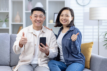 Smiling couple sitting on couch giving thumbs up and holding a smartphone in a bright living room. Happy and cheerful moment. - Powered by Adobe