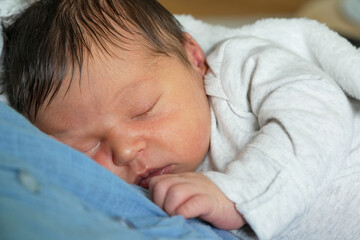 Close-up of a newborn baby sleeping peacefully on mother's chest, wrapped in a white blanket. The...