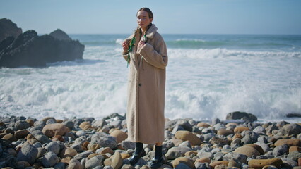 Young woman standing shore in cozy coat. Foaming ocean waves crash rocky coast