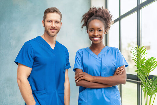 Ein junger Mann mit kurzen Haaren und eine junge Frau mit Afrolook stehen als diverse Pflegefachkr&auml;fte in blauer Arbeitskleidung nebeneinander und l&auml;cheln