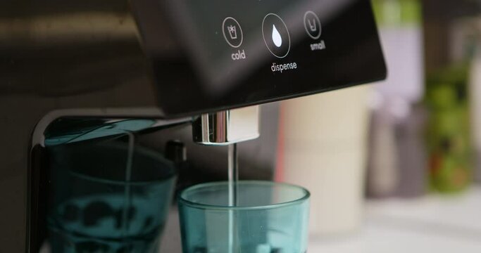 A man in a checkered shirt uses a water filtration machine to fill a blue glass in a contemporary kitchen.