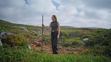 Hiking girl posing mountain hill with stick. Fashion adventurer standing green