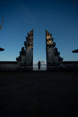 A person standing between two traditional Balinese split gates at dusk.