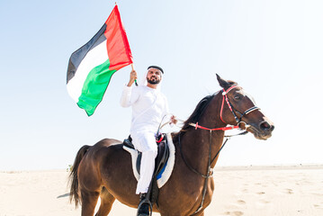 Young adult with Kandura, the emirates traditional clothes, riding his horse in the desert and waving the Uae flag.