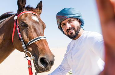Young adult with Kandura, the emirates traditional clothes, riding his horse in the desert and...