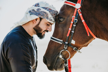 Young adult with Kandura, the emirates traditional clothes, riding his horse in the desert. Tender moment between them staying head against head.