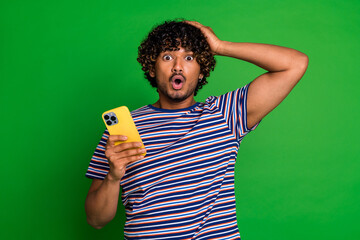 Portrait of curly haired guy in striped t shirt using smartphone touch head confused with internet...