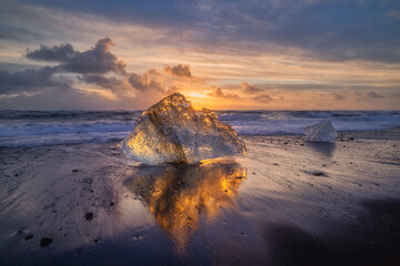 Reflection of an Ice fragment on black sand beach illuminated orange by a setting sun in Iceland
