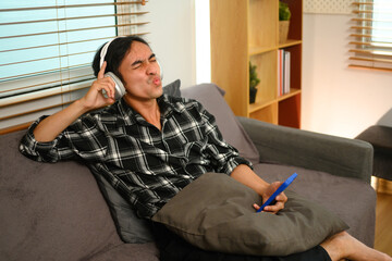 Cheerful asian guy holding mobile phone enjoying favorite songs while relaxing on sofa at home