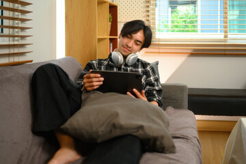 Young man relaxing on couch in living room and using digital tablet for internet surfing