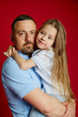 Portrait of young man holds on hands his beloved little daughter against vibrant red studio...