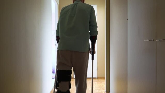 Middle-aged man with leg injury walking on crutches at home. Man wearing leg brace knee support adjustable strap fracture fixator