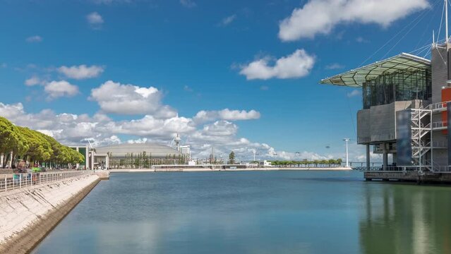 Panorama showing Lisbon Oceanarium timelapse, located in the Park of Nations or Parque das Nacoes. The largest indoor aquarium in Europe. Waterfront with green trees and clouds on a blue sky. Portugal