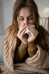 Wrapped in a blanket, a woman sits on the couch at home, fighting a seasonal cold. With a tissue in...