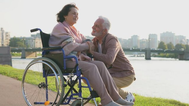 Senior woman in wheelchair walking with caregiver old man on road. Elderly family couple man supporting taking care of paralyzed woman in chair for people with disability outdoor. Rehabilitation